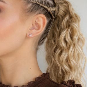 May include: A silver-toned stud earring featuring two connected four-pointed star shapes. The earring is positioned on the earlobe, showcasing a metallic finish and a delicate, angular design that creates a distinct geometric appearance on the skin.