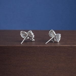 May include: A pair of silver-colored bow-shaped stud earrings. The earrings are on a brown surface with a blue background. The bows have a textured appearance, and the earring posts are visible.