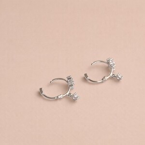 May include: A pair of silver hoop earrings with a cluster of small, square-cut gemstones. The earrings are open and have a delicate, minimalist design. The background is a solid, light pink color.