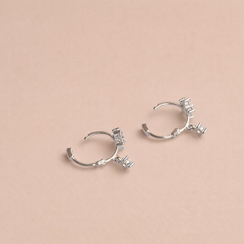 May include: A pair of silver hoop earrings with a cluster of small, square-cut gemstones. The earrings are open and have a delicate, minimalist design. The background is a solid, light pink color.