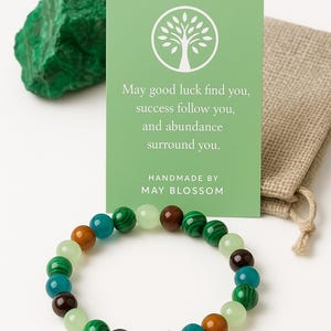 May include: A beaded bracelet with various colored stones, including green, brown, and teal, is displayed with a green card that reads "May good luck find you, success follow you, and abundance surround you." A green stone and a small burlap bag are also in the image.