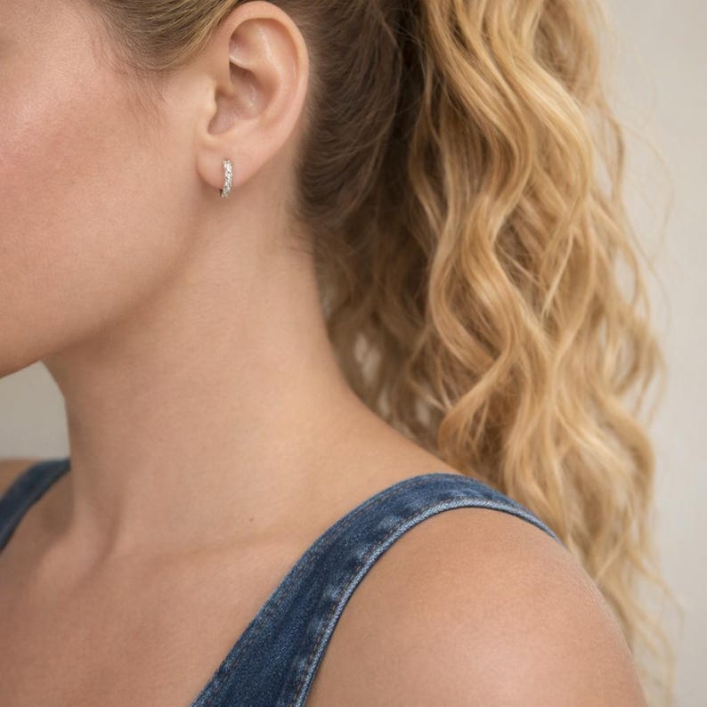 May include: A close-up of a silver hoop earring with small, clear stones. The earring is worn on a person's ear. The person is wearing a denim top, and their blonde hair is pulled back in a ponytail.