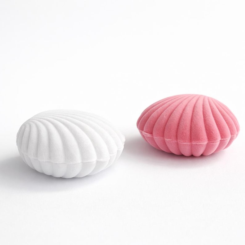 Puede incluir: Dos joyeros en forma de concha. Uno es blanco, el otro es rosa. Ambas cajas tienen una textura suave y aterciopelada y un dise&ntilde;o acanalado, que se asemeja a una concha. Las cajas est&aacute;n sobre un fondo blanco.