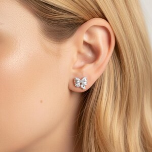 May include: A close-up of a bow-shaped stud earring. The earring is silver-toned and encrusted with multiple small, clear gemstones. The earring is worn on a person's ear, with blonde hair visible.