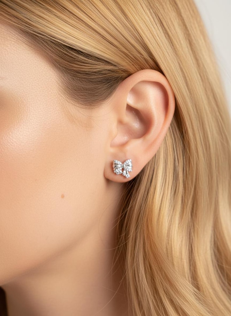 May include: A close-up of a bow-shaped stud earring. The earring is silver-toned and encrusted with multiple small, clear gemstones. The earring is worn on a person's ear, with blonde hair visible.