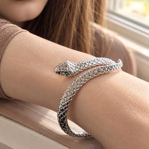 May include: A silver-toned metal bracelet shaped like a coiled snake with a textured, scale-like surface pattern. The snake head features a small, vibrant green gemstone set as an eye, and the body wraps around the arm in a spiral design.