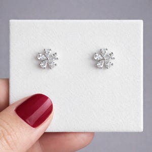 以下が含まれることがあります： 銀色のスタッドピアス2つ。それぞれに花の模様があります。花は、中心点を取り囲むように配置された、6つの透明な洋梨型の宝石で構成されています。ピアスは白い表面に表示されています。