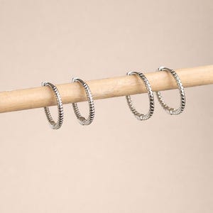 May include: Four silver hoop earrings with a textured, ridged design are displayed on a wooden dowel against a neutral background. The earrings are evenly spaced and appear to be made of metal.
