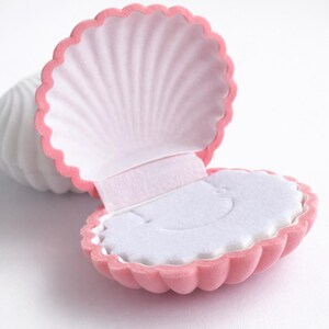Puede incluir: Una caja de joyer&iacute;a en forma de concha rosa, abierta para revelar un interior blanco. La caja tiene un borde festoneado y una textura suave y aterciopelada. El interior est&aacute; forrado con tela blanca, proporcionando un delicado contraste con el exterior rosa.