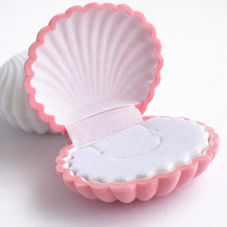 Puede incluir: Una caja de joyer&iacute;a en forma de concha rosa, abierta para revelar un interior blanco. La caja tiene un borde festoneado y una textura suave y aterciopelada. El interior est&aacute; forrado con tela blanca, proporcionando un delicado contraste con el exterior rosa.