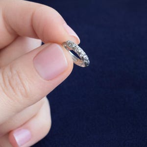 May include: A silver ring with a band of small, clear gemstones. The ring is held against a dark blue background. The gemstones are set closely together, creating a sparkling effect. The ring appears to be a wedding or anniversary band.