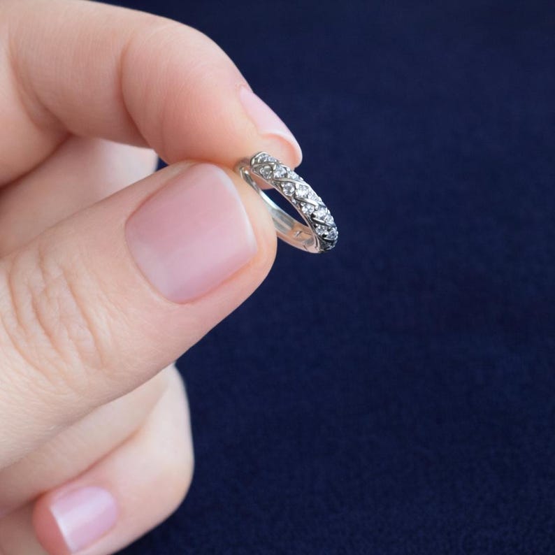 May include: A silver ring with a band of small, clear gemstones. The ring is held against a dark blue background. The gemstones are set closely together, creating a sparkling effect. The ring appears to be a wedding or anniversary band.