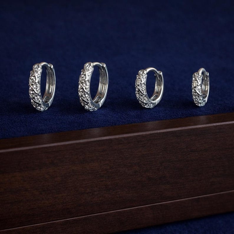 May include: Four silver hoop earrings with a textured design and small, clear stones. The earrings are displayed on a dark blue surface, with a dark brown wooden box in the foreground. The earrings vary in size.