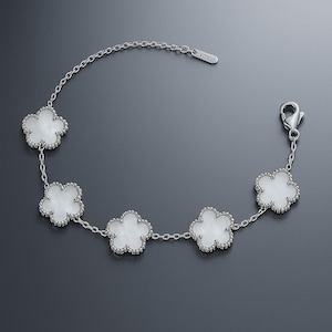 Op de afbeelding: Een zilveren armband met vijf witte klavervormige bedels. Elke bedel heeft een kralenrand en is verbonden door een fijne ketting. De armband heeft een karabijnsluiting.