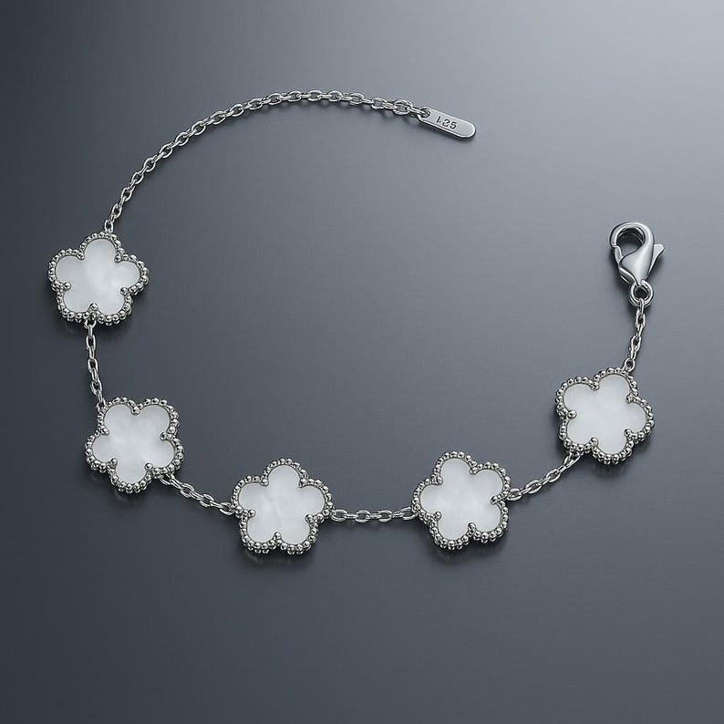 Op de afbeelding: Een zilveren armband met vijf witte klavervormige bedels. Elke bedel heeft een kralenrand en is verbonden door een fijne ketting. De armband heeft een karabijnsluiting.