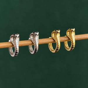 May include: Two pairs of hoop earrings shaped like cobra snakes, with one pair in a polished silver tone and the other pair in a polished gold tone. Each earring features a detailed scale texture along the body and a raised hood design.