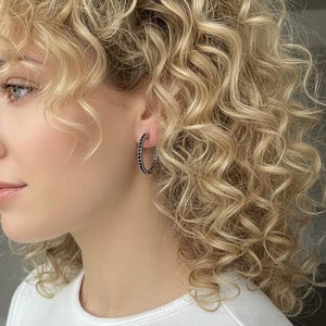 以下が含まれることがあります： カールしたブロンドの髪の毛を持つ人が、白いトップスと黒いフープイヤリングを身に着けているクローズアップ。イヤリングには小さな暗い石がちりばめられています。イヤリングと髪の毛に焦点が当てられています。