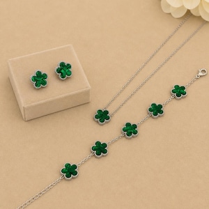 Può includere: Un set di gioielli composto da collana, bracciale e orecchini. Ogni pezzo presenta una montatura color argento con un design a quadrifoglio di colore verde brillante. Gli orecchini sono esposti in una piccola scatola quadrata beige.