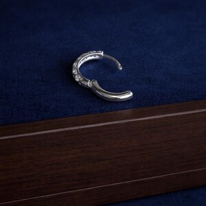 May include: A silver hoop earring with small, clear gemstones set along the outer curve. The earring is open, revealing the clasp mechanism. The background is a dark blue fabric, and a wooden frame is visible at the bottom.