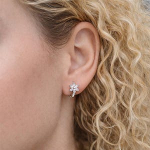 May include: A silver-toned hoop earring featuring a flower-shaped charm adorned with six clear, marquise-cut stones arranged in a petal pattern. The earring is secured through the earlobe, showcasing a metallic finish and sparkling white stones.