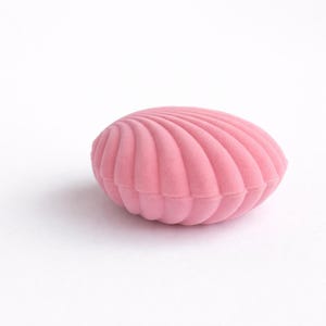 Puede incluir: Una caja de joyer&iacute;a rosa en forma de concha con una textura suave y aterciopelada. La caja es de un tono rosa claro y tiene un dise&ntilde;o acanalado, que se asemeja a una concha. La caja est&aacute; cerrada y sobre una superficie blanca.