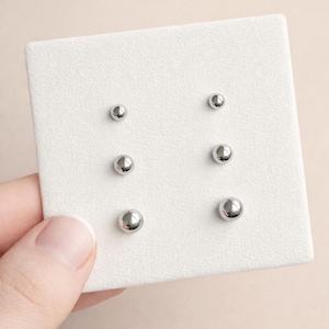 Puede incluir: Seis pequeños pendientes de botón esféricos plateados y brillantes están dispuestos sobre una tarjeta de exhibición blanca texturizada. Los pendientes están en tres pares de diferentes tamaños. La tarjeta la sostiene una mano de piel clara.