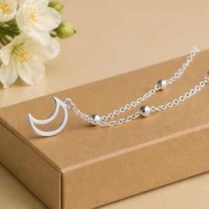 May include: A silver-coloured necklace featuring a crescent moon pendant attached to a delicate chain. The chain is accented with small, polished silver-coloured spherical beads spaced evenly along the links, creating a simple and reflective design.