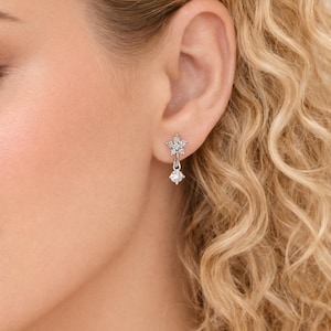 May include: A close-up of a woman's ear adorned with a silver earring. The earring features a star-shaped top with a dangling round gem. The jewelry sparkles, reflecting light. The woman has blonde, curly hair.