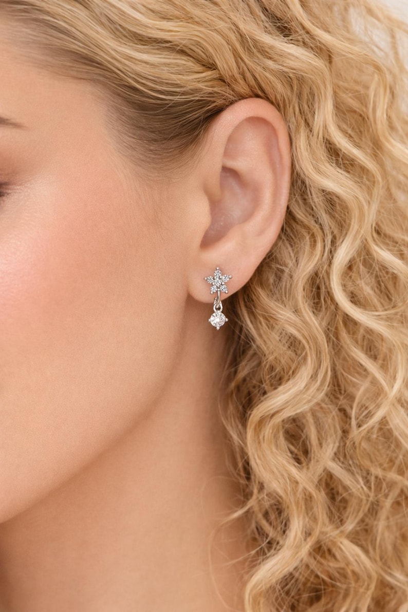 May include: A close-up of a woman's ear adorned with a silver earring. The earring features a star-shaped top with a dangling round gem. The jewelry sparkles, reflecting light. The woman has blonde, curly hair.