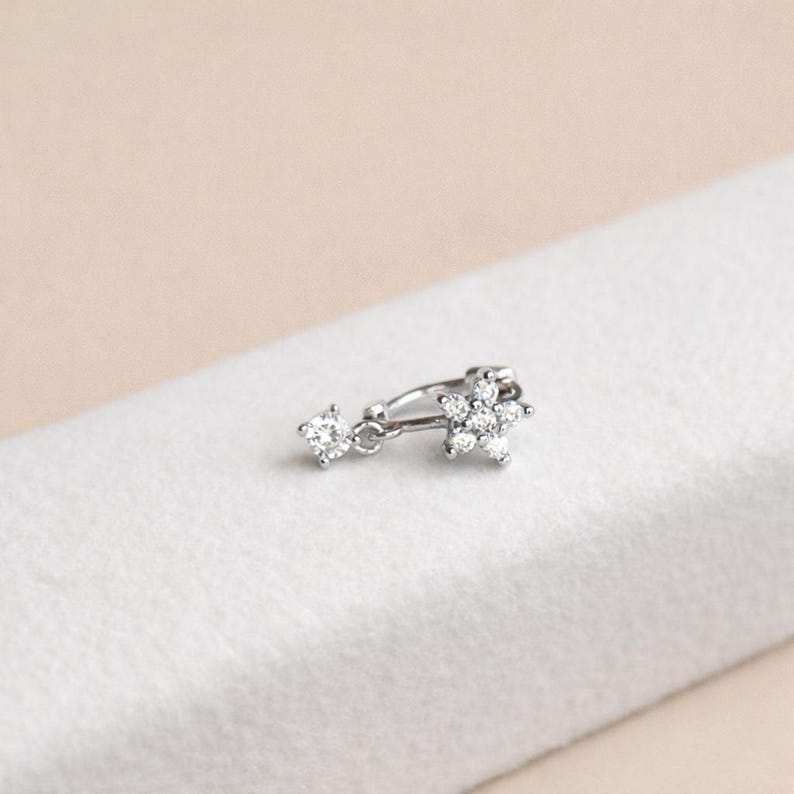 May include: A silver-toned earring featuring a dangling, round-cut gemstone and a flower-shaped cluster of gemstones. The earring is set against a white background, with a soft, neutral backdrop.