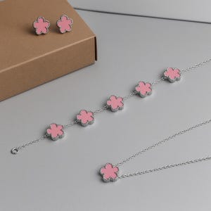 May include: A jewellery set comprising a necklace, bracelet, and earrings. Each item features a silver-coloured metal flower design with a pink centre. The earrings are studs, and the bracelet and necklace have multiple flowers.