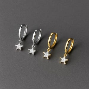 May include: Four hoop earrings with star-shaped charms. Two earrings are silver-toned, and two are gold-toned. Each earring features a small, sparkling star pendant suspended from a hinged hoop. The earrings are displayed against a dark gray background.