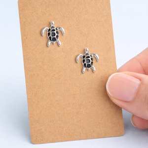 May include: A pair of silver-coloured turtle stud earrings with black detailing, presented on a brown card. The earrings are small and detailed, with the turtle's shell and limbs visible. The card is held by a hand.