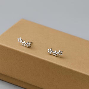 Può includere: Un paio di orecchini a forma di stella color argento. Ogni orecchino presenta tre stelle in fila, con un perno per l'inserimento. Gli orecchini sono esposti su una superficie marrone.