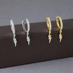 May include: Two pairs of hoop earrings. One pair is silver-toned, the other is gold-toned. Both pairs feature a row of small, clear stones along the hoop. Each earring has a lightning bolt charm dangling from the bottom, also adorned with clear stones.