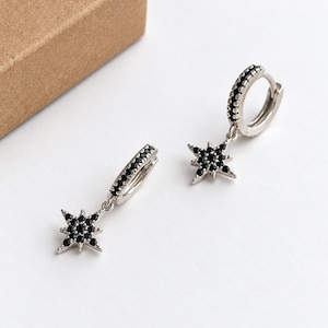 May include: A pair of silver hoop earrings featuring a dangling star-shaped charm. Each hoop and star is encrusted with small, round black stones. The earrings have a hinged closure mechanism and a polished metallic finish throughout the design.