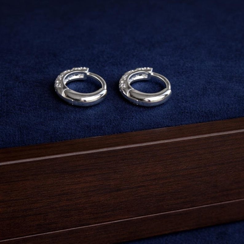 May include: Two silver hoop earrings with small, clear stones set along the top curve. The earrings are displayed on a dark blue velvet surface, with a dark wood border at the bottom of the image.