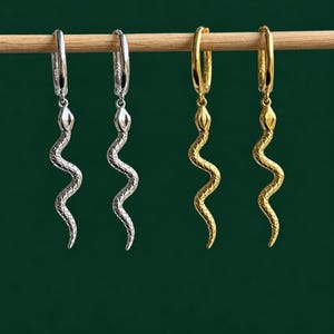 Puede incluir: Cuatro pendientes de aro colgados de una varilla de madera. Dos pendientes son de tono plateado y dos de tono dorado. Cada pendiente presenta un dije de serpiente con un patrón texturizado de escamas en el cuerpo y una cabeza definida.