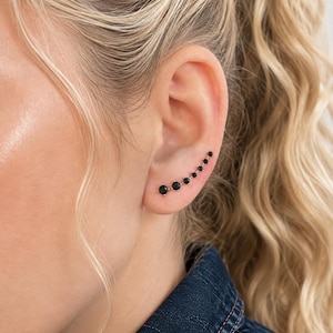 Op de afbeelding: Een ear climber oorbel met een rij van zeven ronde zwarte stenen in een metalen zetting. De stenen worden geleidelijk kleiner naarmate ze de curve van de oorlel naar boven volgen, wat een lineair ontwerp creëert dat langs de buitenrand zit.