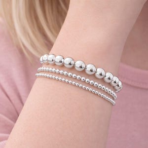 Puede incluir: Un conjunto de tres pulseras apiladas con cuentas esféricas de tono plateado pulido. La pulsera superior consta de cuentas grandes, mientras que las pulseras central e inferior están compuestas por esferas metálicas uniformes.