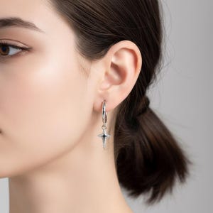 以下が含まれることがあります： スターバーストペンダント付きのシルバーフープピアス。スターバーストペンダントは黒い中心部を持ち、小さな輝くクリスタルで縁取られています。ピアスは人の耳に装着されています。
