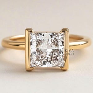May include: A gold ring with a square, clear gemstone. The gemstone is set in a bezel setting with vertical gold bars. The ring band is a simple, rounded gold band. The text "KIVA JEWEL" is visible on the side of the ring.