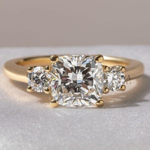 Peut inclure: Une bague en or avec une grosse pierre centrale en diamant de taille carrée, flanquée de deux diamants ronds plus petits. La bague a un anneau simple et poli et est posée sur un fond doux et neutre. La bague est un bijou.