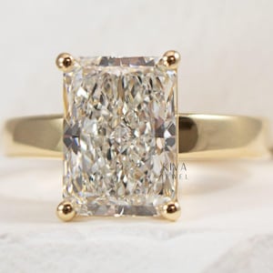 May include: A radiant-cut diamond ring with a gold band. The rectangular diamond is set with four gold prongs. The ring is set on a white surface. The text "KIVA JEWEL" is visible on the right side of the image.