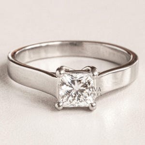 May include: A silver ring featuring a square-cut diamond. The ring has a minimalist design with a smooth band and four prongs holding the diamond. The ring is set against a white backdrop.