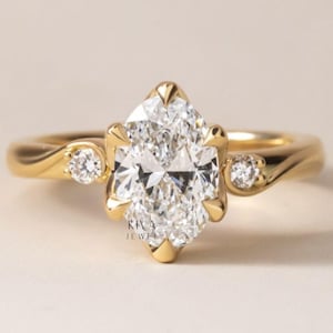 May include: A gold engagement ring with a large, marquise-cut diamond as the centerpiece. Two smaller diamonds flank the main stone. The ring band has a smooth, flowing design. The text "KIVA JEWEL" is visible.