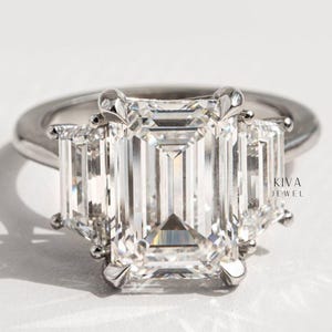 Peut inclure: Une bague en platine ornée d'un grand diamant central de taille émeraude, flanqué de deux diamants de taille émeraude plus petits. La bague présente un design classique à trois pierres. Le nom de la marque "KIVA JEWEL" est visible.