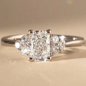 May include: A diamond engagement ring featuring a central rectangular-cut diamond, complemented by two clusters of smaller diamonds. The ring is set in a polished silver band. The design highlights the sparkle of the gemstones, a classic piece of jewellery.