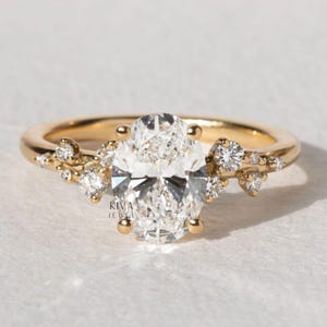 May include: A gold ring with a large, oval-cut diamond as the focal point. Smaller diamonds are set on either side of the main stone, forming a delicate, nature-inspired pattern. The ring has a polished, yellow gold band.