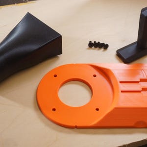 May include: An assortment of black and orange 3D-printed parts, likely for a woodworking tool. The components include a black nozzle, an orange base with a circular opening, a black handle, and small black screws. The parts are arranged on a light-colored wooden surface.